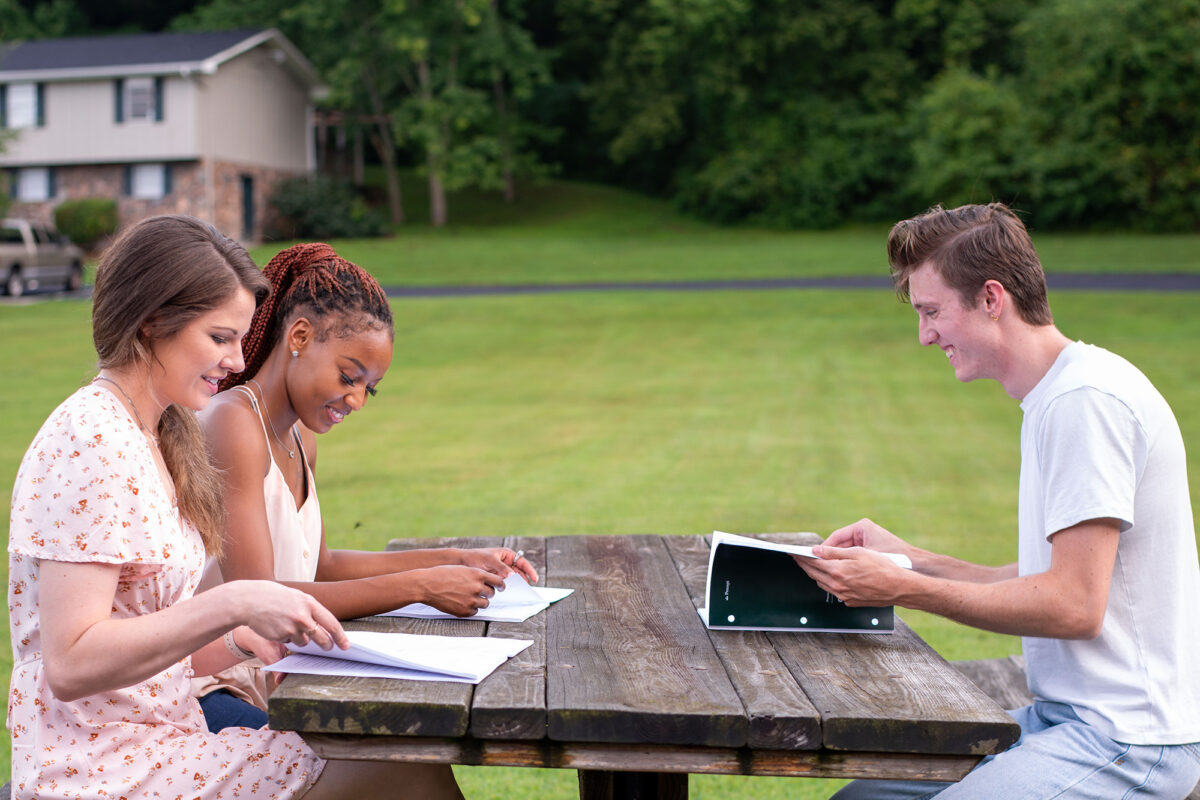 Three young people sitting at a picnic table outside discussing a Precept Bible study together, applying God's word to your life